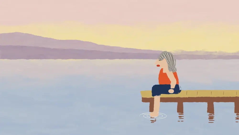 A woman with grey hair sitting alone at the end of a jetty, one foot in the lake.